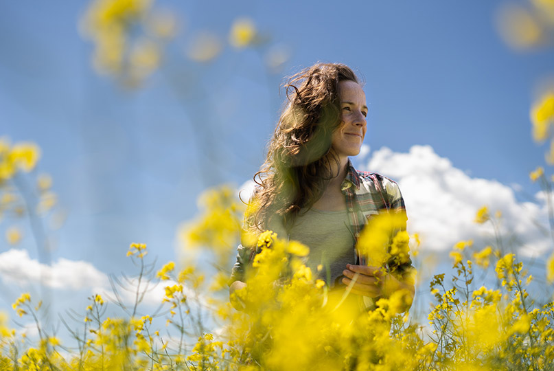 Mujer sonriente de pie en medio de una granja de colza, con un cielo azul claro y hermoso de fondo.