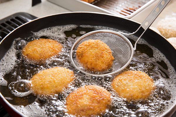 Breaded items being deep-fried in oil in a frying pan, with a metal slotted spoon handling them.