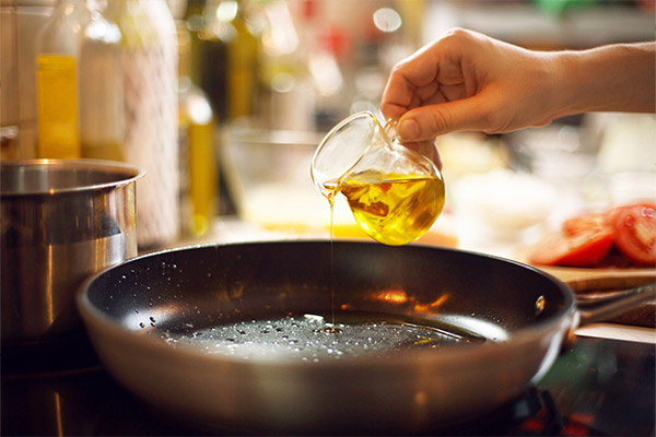 A person’s hand pouring oil from a small glass jug into a heated frying pan on a stove. The pan has bubbles forming, indicating it is hot, and blurred kitchen utensils and tomatoes are visible in the background.