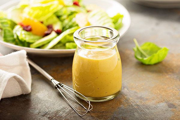 A glass jar of yellow dressing next to a fresh salad and a metal whisk on a table.