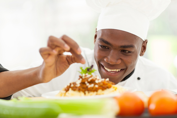 Smiling chef garnishing a plate of pasta with herbs, surrounded by fresh vegetables in the foreground.