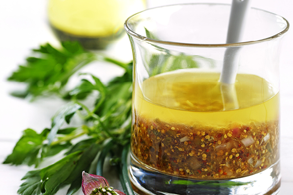 Glass jar filled with a vinaigrette made from mustard seeds and oil, with fresh parsley in the background. A white spoon rests in the jar.